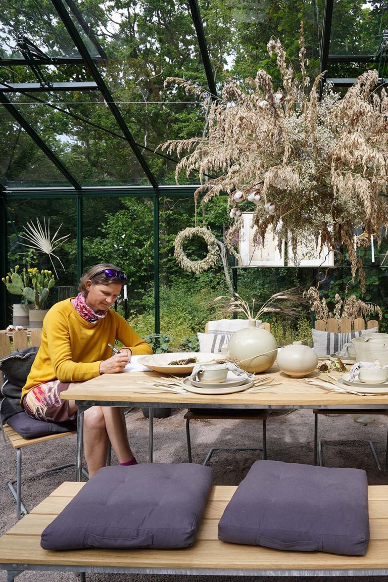 Für Schweden typisches Glashaus mit Dekoration. Die Bloggerin Judith Beck sitzt an einem Tisch im Glashaus und schreibt in ein Buch. Boho-Deko ist im Raum verteilt.