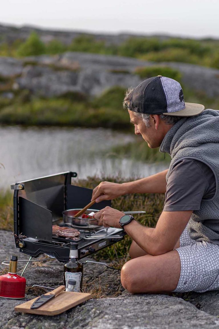 Der Blogger Jesús Tena sitzt auf grauen Steinen am Rand eines Sees in Schweden. Er hat einen Campinggrill von Primus und bereitet darauf frischen Lachs und Gemüse zu.
