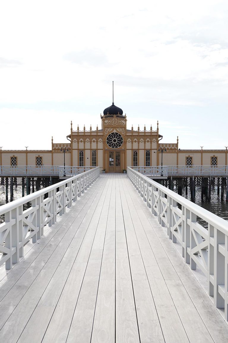 Eines der schönsten Kaltbadehäuser an der Westküste in Schweden mit einem langen Zugangssteg in gebrochenem Weiß und dem historischen Gebäude im Hintergrund.