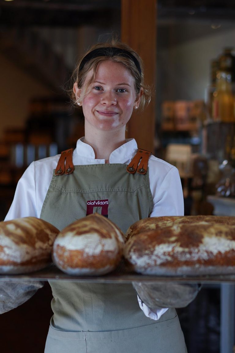 In einer Stenugnsbageri in Schweden hat eine junge Mitarbeiterin frisches Brot aus dem Ofen geholt und präsentiert 3 Laibe auf einem Tablett.