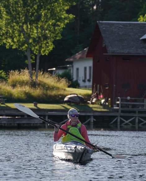 Die Bloggerin Judith Beck vom Fernzeit Reiseblog paddelt auf dem Dalsland Kanal mit dem Kajak. Es ist Sonnenuntergang und das Licht ist kontrastreich. Dahinter steht ein typisches rotes Schwedenhaus.